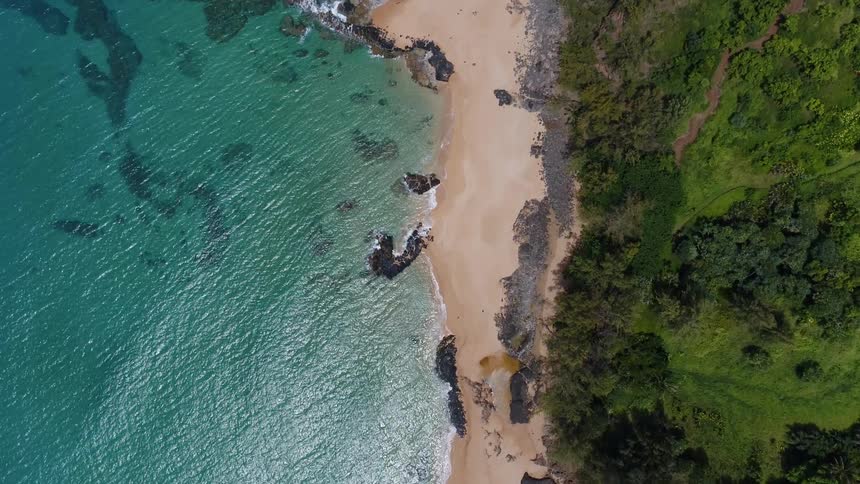 Waves from a clear blue ocean lap a sandy beach that is overlooked by hills covered in vegetation