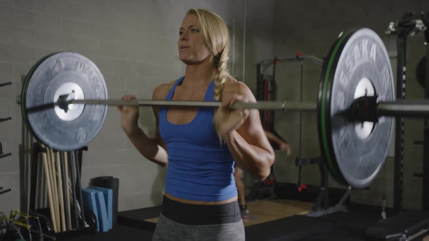 Muscular woman lifts barbell in a gym with people working out in the background during daylight