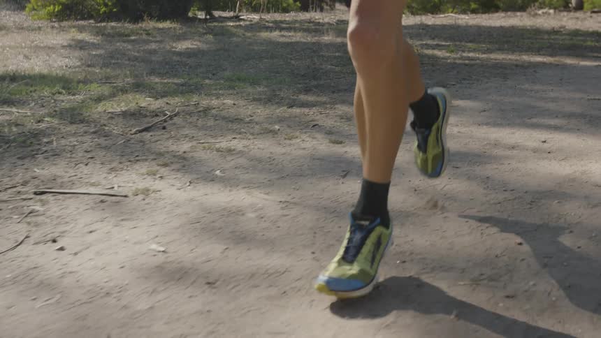 A man in black shorts and colorful running shoes is jogging on a dirt path in a park