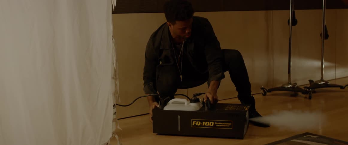 A man smooths out a curtain and then crouches down to turn on a fog machine inside of a darkened room