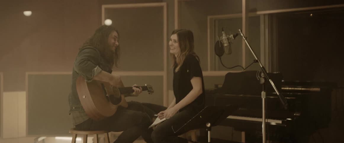 A man strums on an acoustic guitar as his female companion sings as both are sitting next to a black piano in a music studio