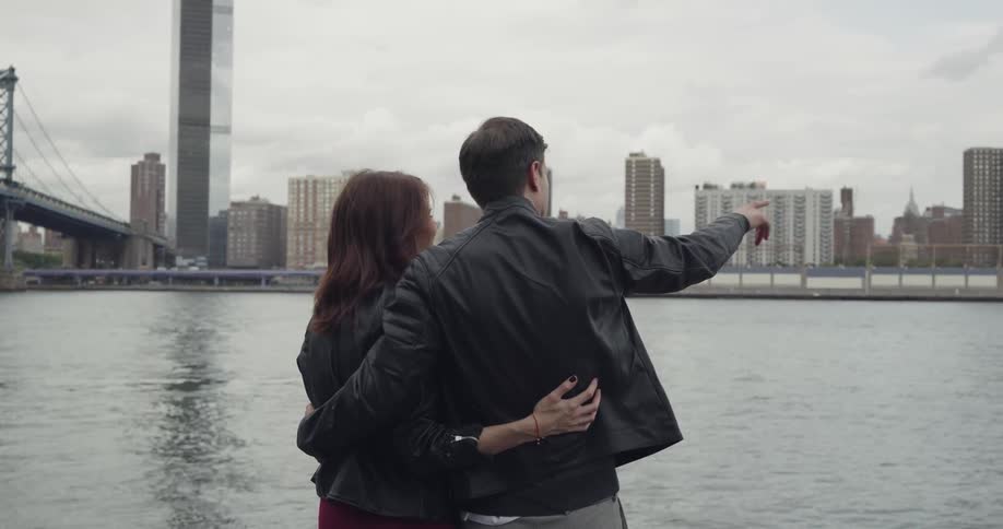 A man and woman hug while looking at a city bridge and distant skyscrapers next to a river