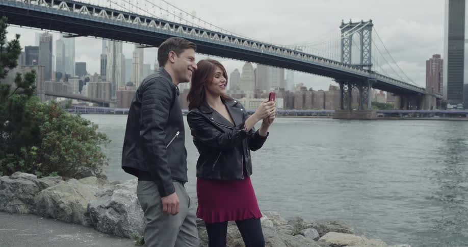 A man and a woman take selfies standing on the waters edge outside of new york city
