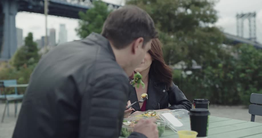 A man and woman both wear leather jackets and sit and eat lunch by a bridge