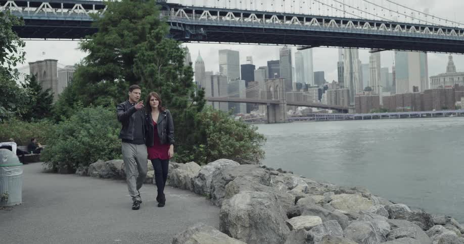 A couple leans into each other as they walk on a park path near the harbor bridge and city skyline