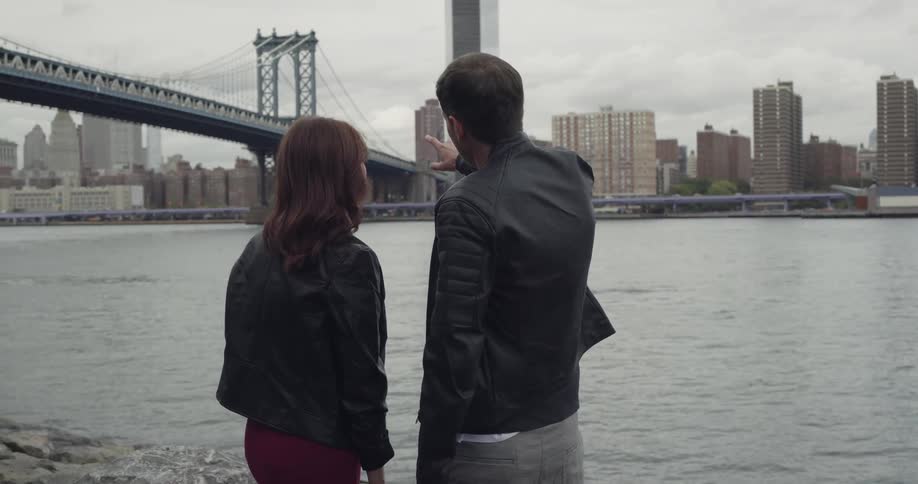 Man and woman standing at riverbank outside a city