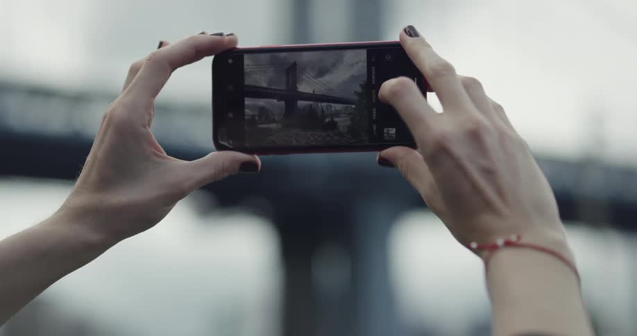 A woman with painted fingernails takes a photo of a city bridge with a smartphone