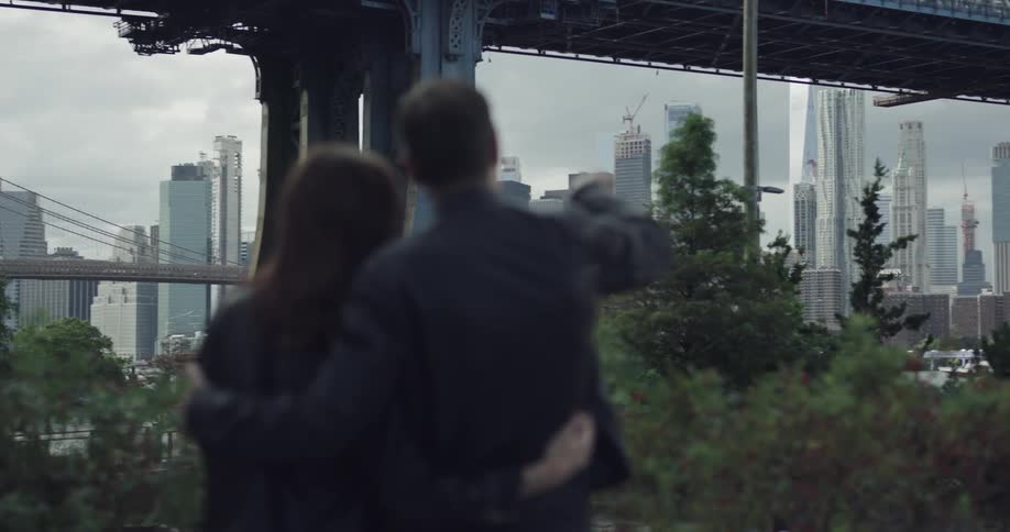 A woman and man in leather jackets embracing one another while looking out and gesturing at the underside of a massive bridge over a bay