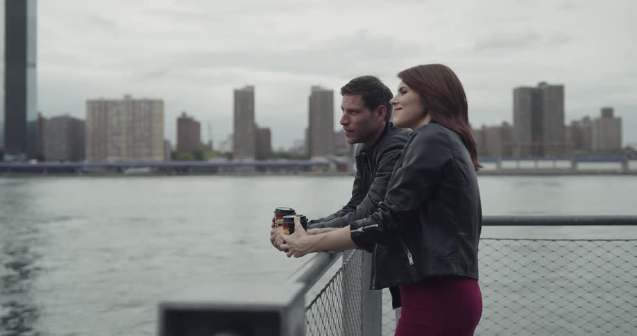 A man and woman in leather jackets holding cups of coffee while leaning on a fence and looking out over the ocean