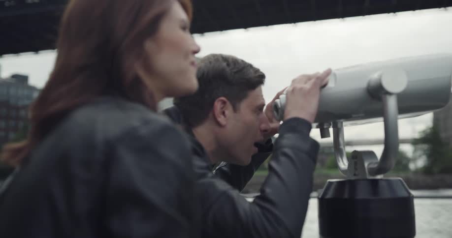 A man and a woman use public binoculars in a park outside of a large city