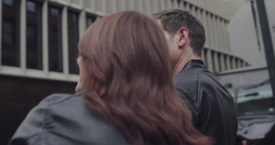A man and woman walk down a sidewalk past businesses in a city