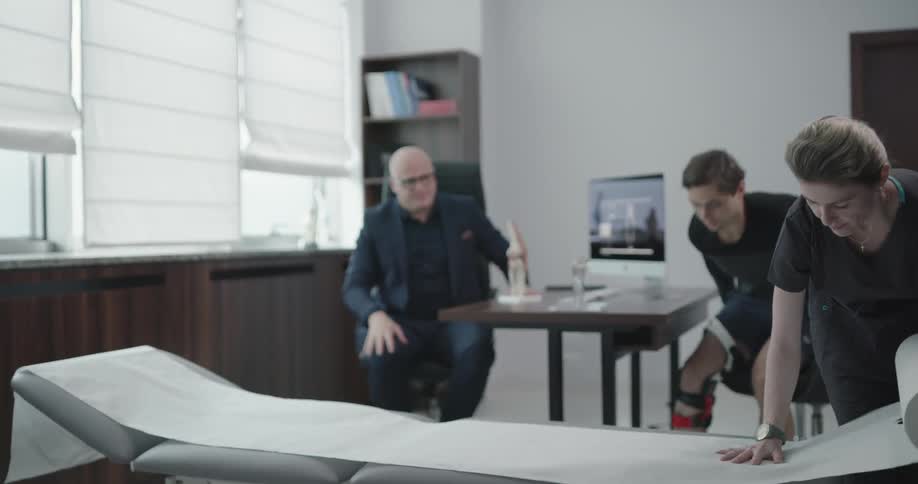One female rolls paper onto a doctor bed while a doctor talks to his patient in a leg brace
