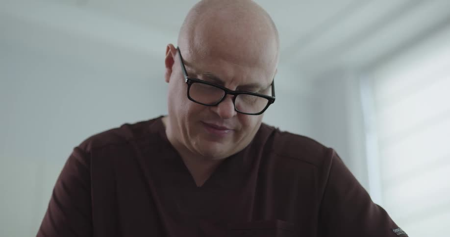 A bald man is looking down at something in a white room and then turns to talk