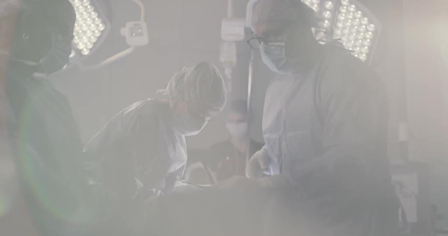 A team of surgeons focus on a patient on their exam table while they work in a well lit surgical suite