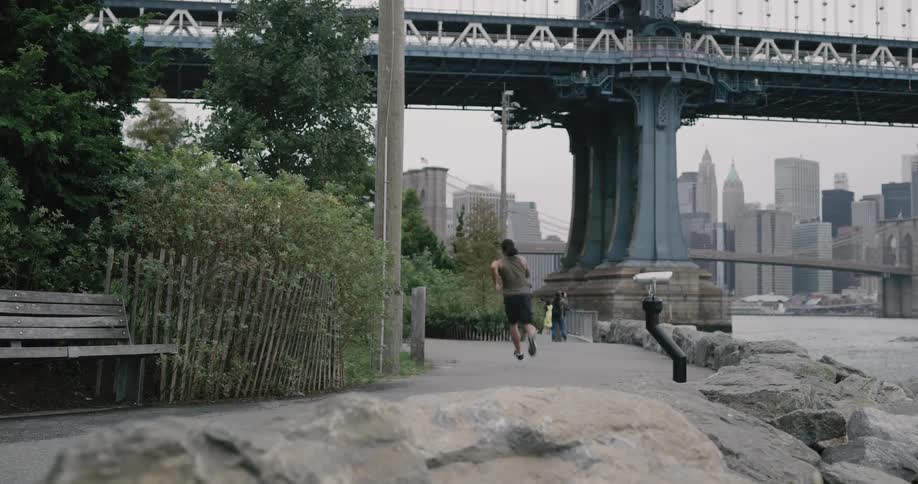 Black man runs along on the promenade next to the river across manhattan bridge and brooklyn bridge on a cloudy day