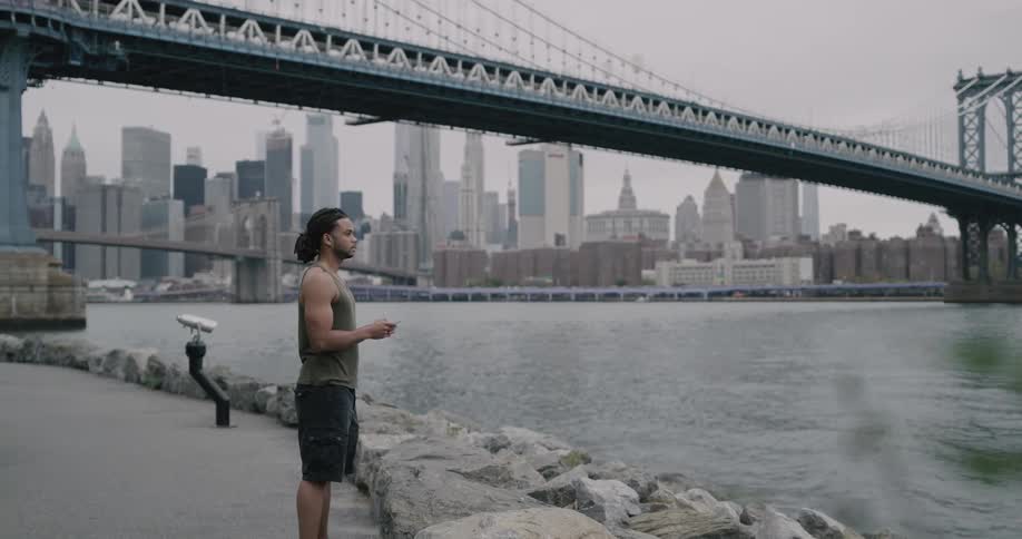 Black man stands and uses smartphone on the promenade next to the sea across the city on a cloudy day