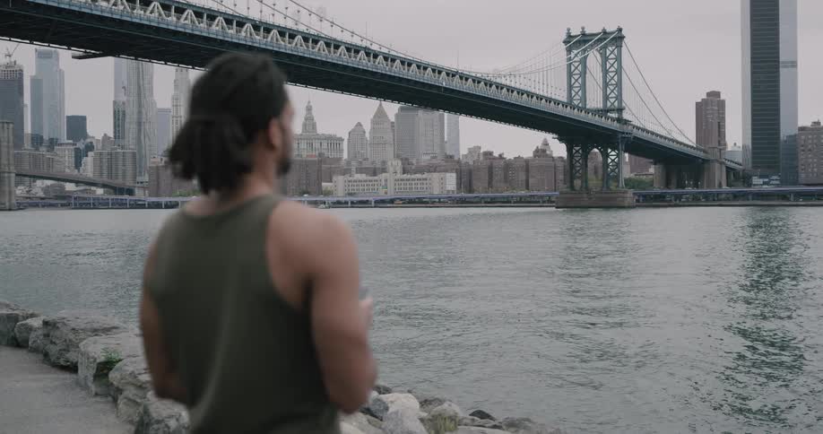A man in a tank top stands next to the water running under a bridge