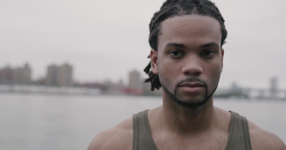 A young black man standing before a bay bordering a city smiling at the camera