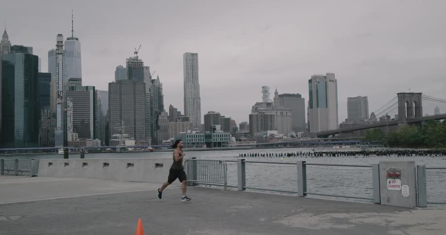 A muscular man runs along a waterfront path near a major metropolitan city as a female jogs by in the opposite direction