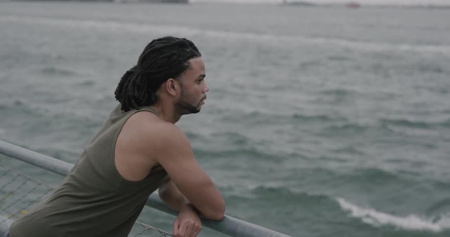 Black man puts elbows on the banister and looks at the sea on a cloudy day