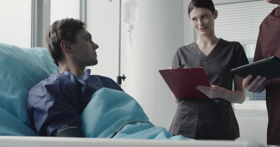 A man and woman holding charts stand at a patients bed and talk with him