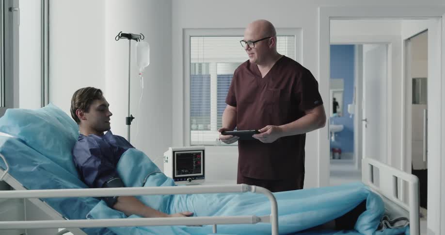 A man walks into a hospital room and shakes the patients hand who is lying in a bed