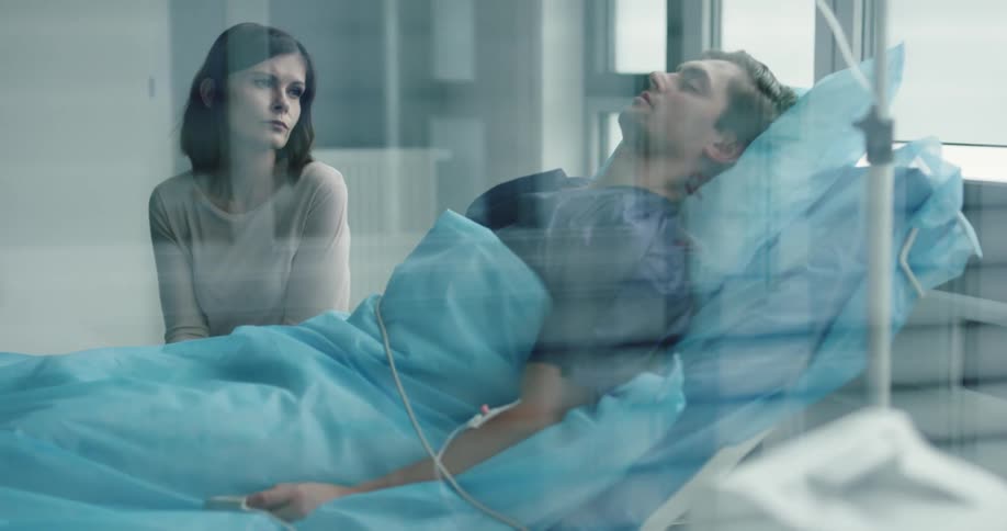 A woman looking worriedly at a man sleeping in a hospital bed