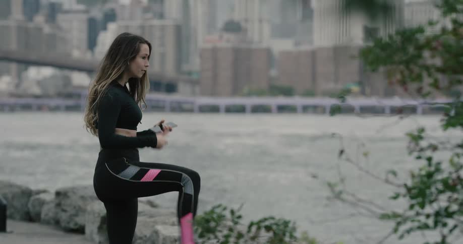 Woman holding smartphones runs by the sea side and takes a break to wear bluetooth earphones across the city