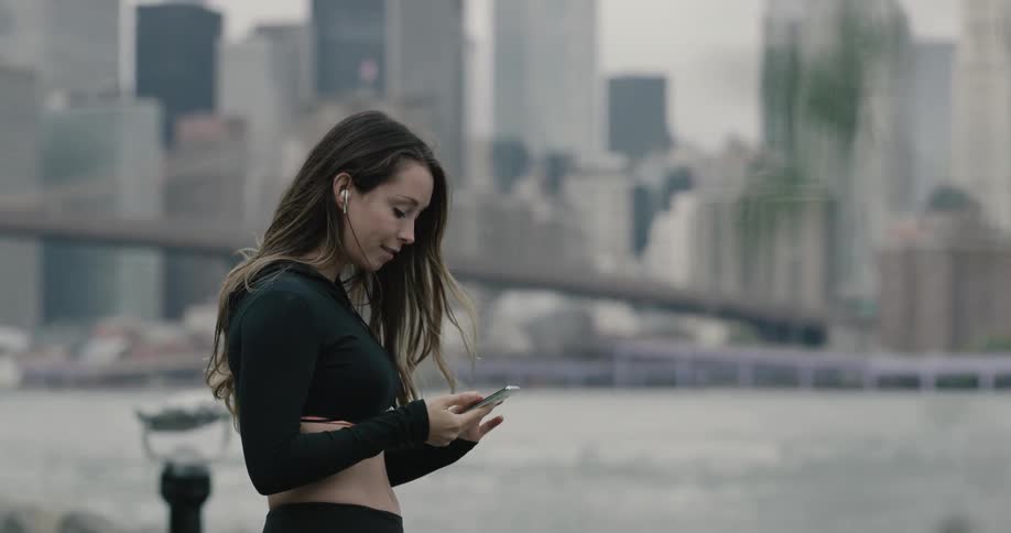 A young woman in athletic clothing listening to music on her smartphone while standing at riverside in a city