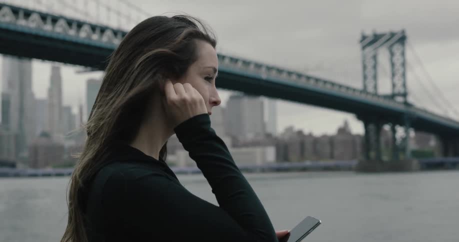 A girl running at the park under the brooklyn bridge stops and ajusts her air pods and glances at her phone