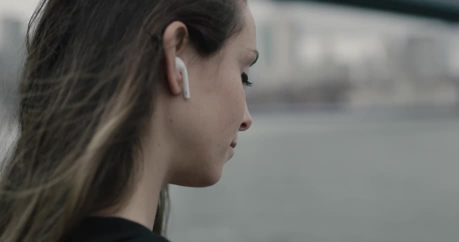 A woman puts a wireless headphone in her right ear as her long hair blows in the wind