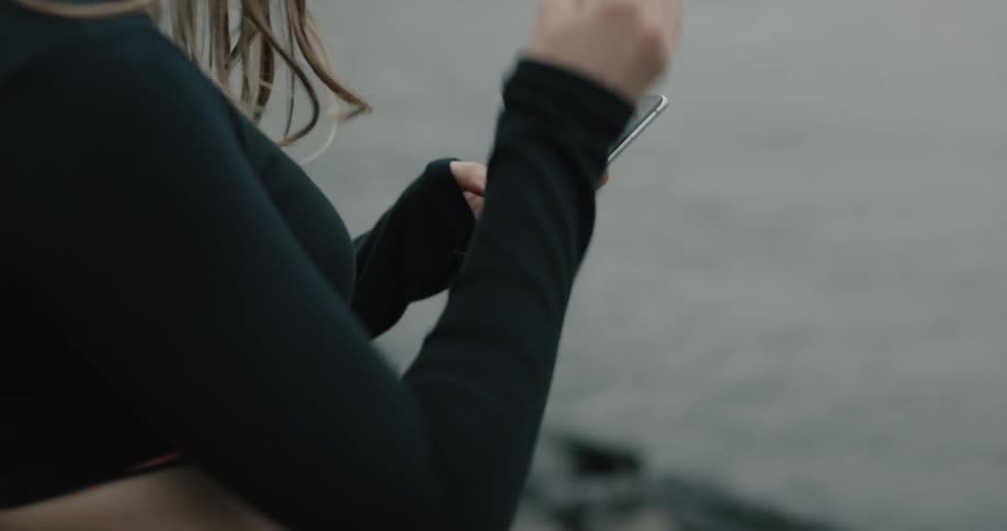A woman puts in headphones while holding a smartphone next to a river in a city