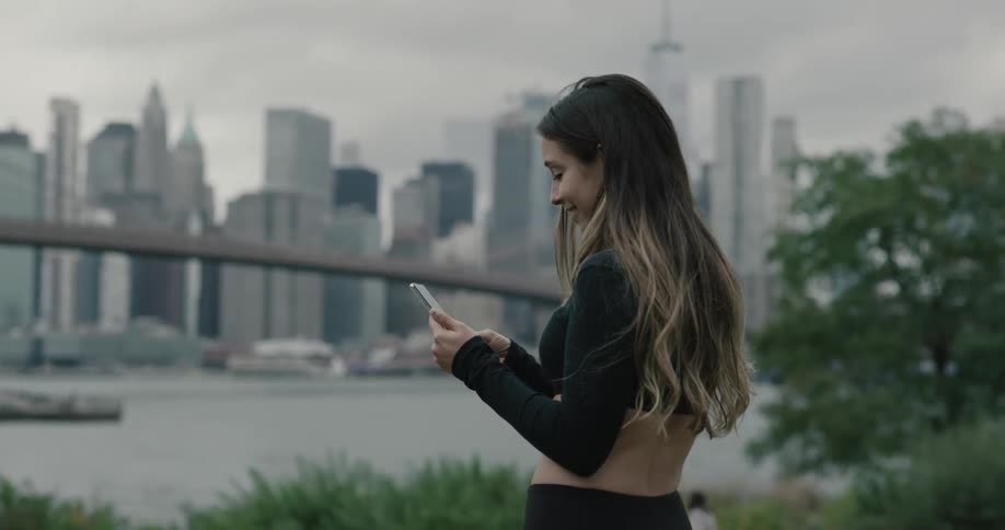 Woman holding smartphone walks to the seaside across the city and smiles during daylight