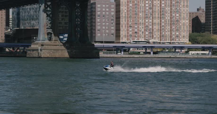 A person on a jet ski is riding along a body of water located next to a metropolitan area as a motorized boat passes by in the other direction