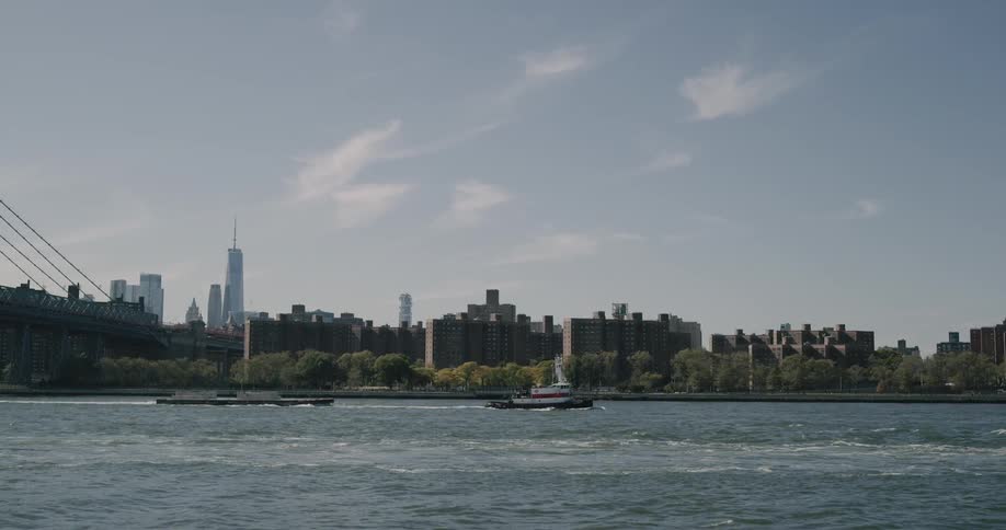 Tug boat pulls barge through water with suspension bridge and city on horizon