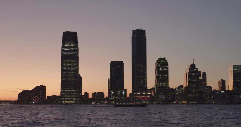 Ferry floats along the sea with skyline of the city with tall buildings next to the sea at dusk