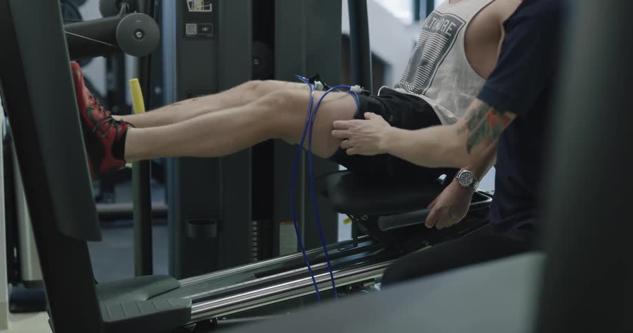 A fit man sits on a piece of fitness equipment and performs some leg press exercise while being encouraged by a fitness trainer