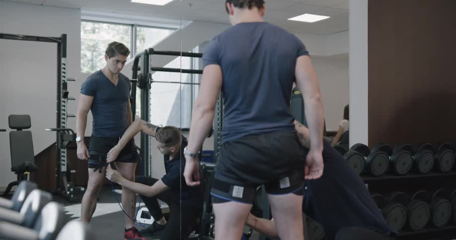 A muscular man stands before a mirror in a fitness room as another man attaches straps around his thighs that are connected by cords to an electric device