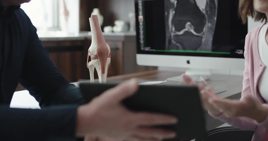 A woman sits in a doctors office as he talks with her and shows her a tablet