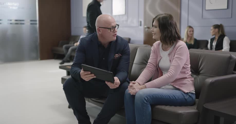 A man holds a tablet and discusses something with a woman in a waiting room