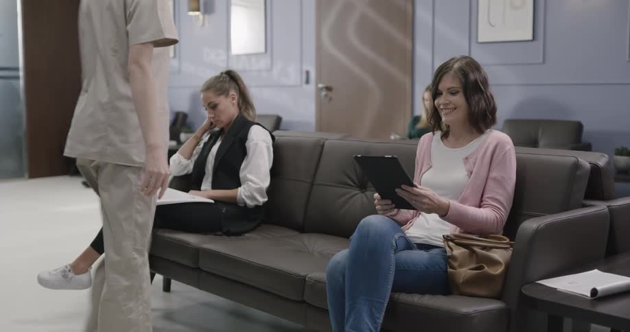 A female medical professional wearing scrubs hands a computer tablet to a female patient and then walks towards a door exiting the waiting room area