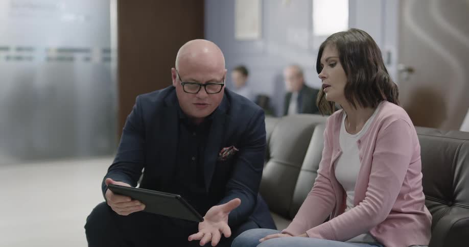 A man sits on a couch and talks with a woman while showing her a tablet