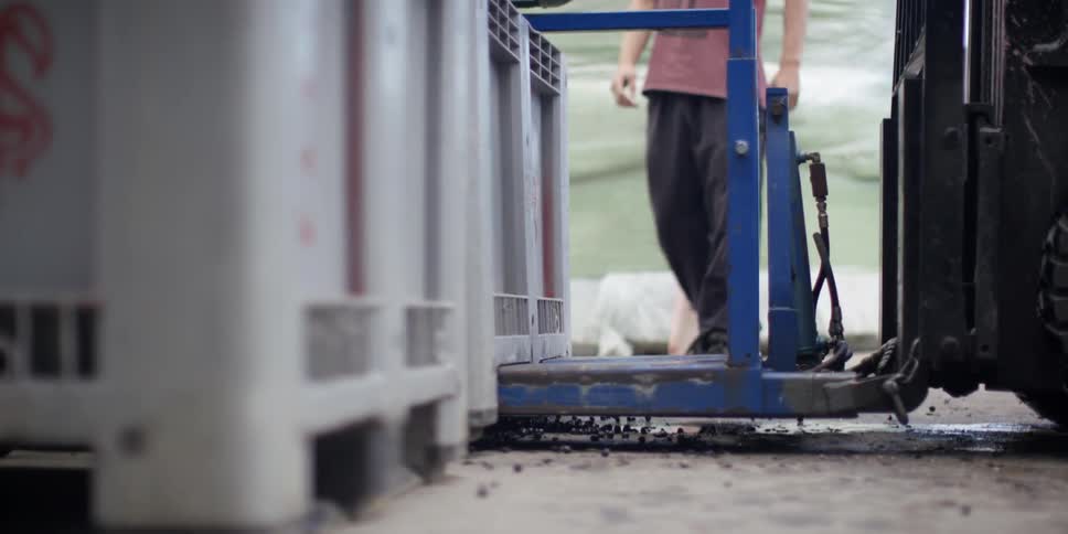 A young man stands near a forklift as it begins lifting a large bin of items