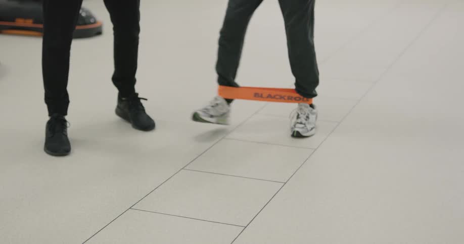 Two males working out while one shows another how to use an orange resistance band