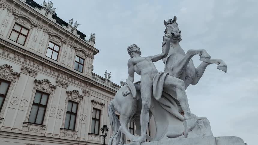 Clouds in the sky move over a statue of a rearing white horse and a man holding on in front of the belvedere palace in vienna
