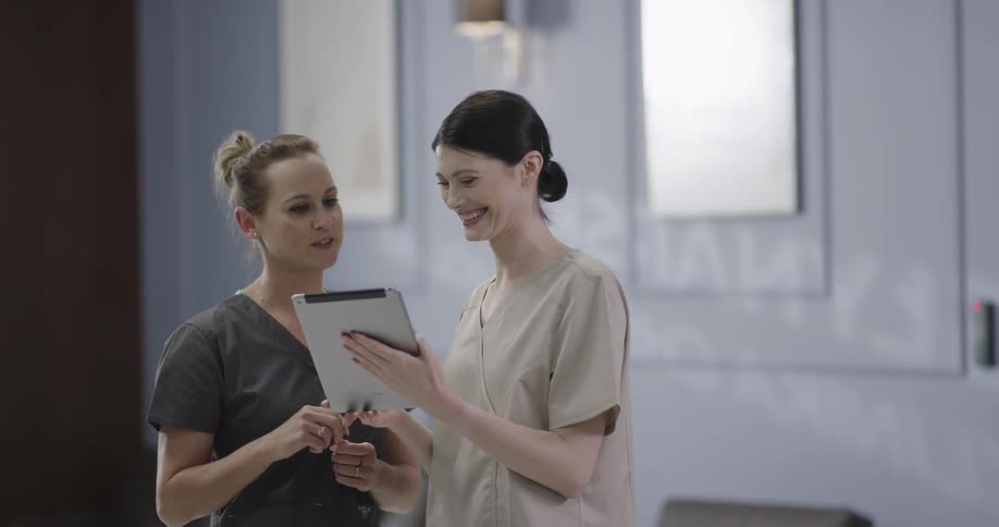 Two female nurses in scrubs smile while looking at a tablet in a clinic
