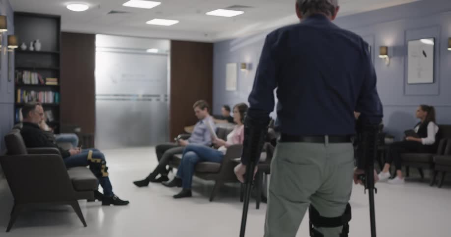 A man with a leg brace is walking with arm canes into a waiting room with several people when a female nurse walks to him and directs him to another place