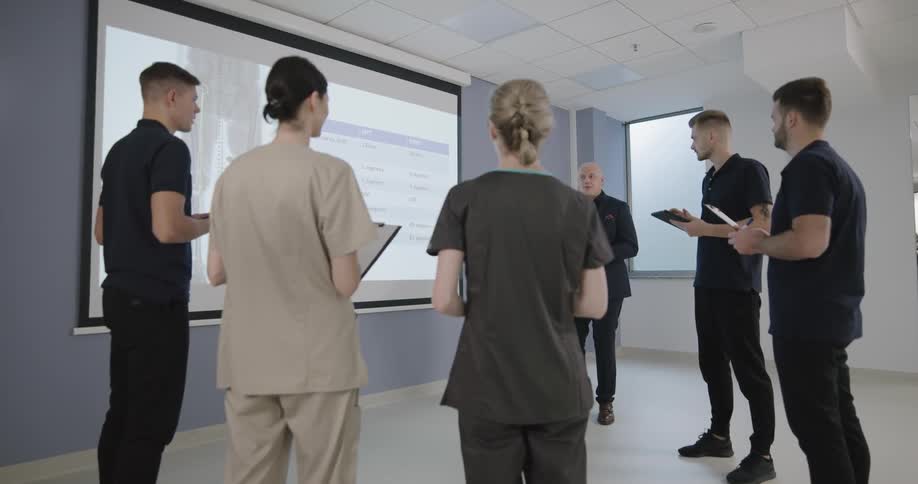 Five people stand holding clipboards as they listen to one person talk as they all look at a screen