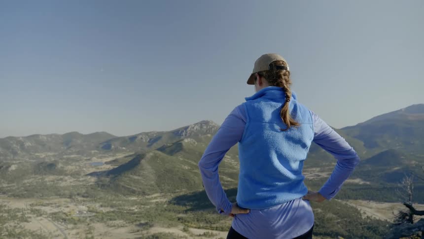 A woman stands on top of a mountain peak and looks down
