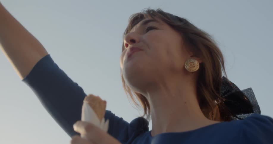 A woman waves while eating an ice cream cone outside on a sunny day